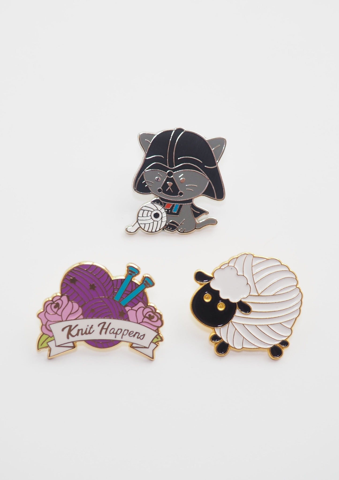 image of three enamel pins, one is a cat in a darth vader costume playing with yarn that looks like the death star, the second is purple balls of yarn with the words "knit happens", and the third is an adorable white sheep with a black face