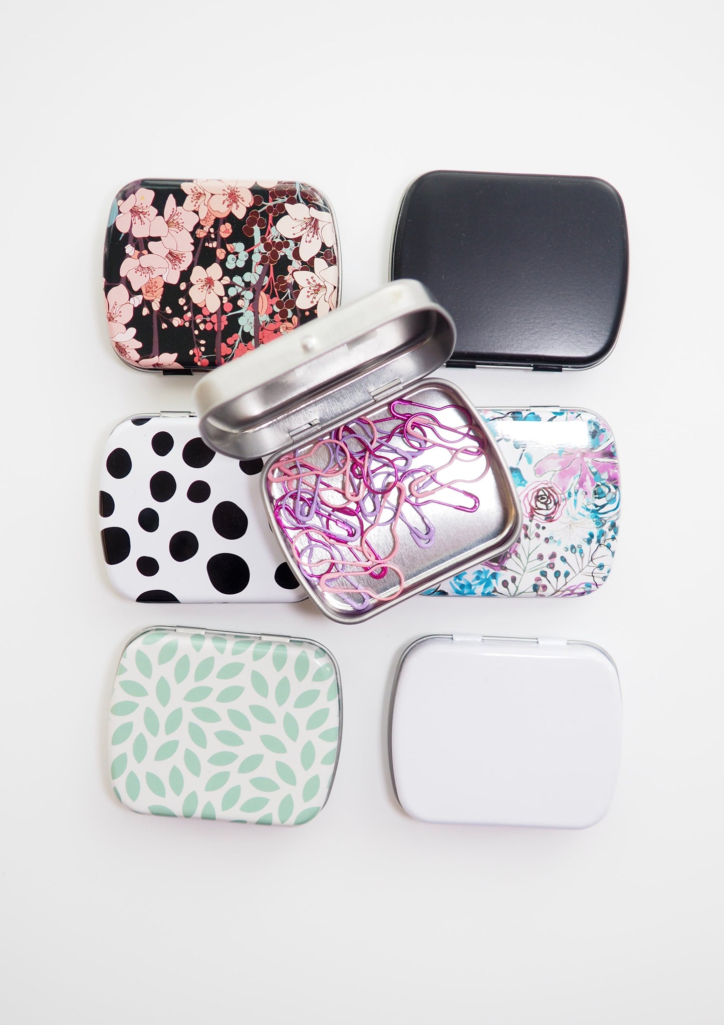 image of metal notion tins. one black with flowers, one with dalmation spots, one white with teal leaves, one blackl, one white, one silver, and one white with flowers