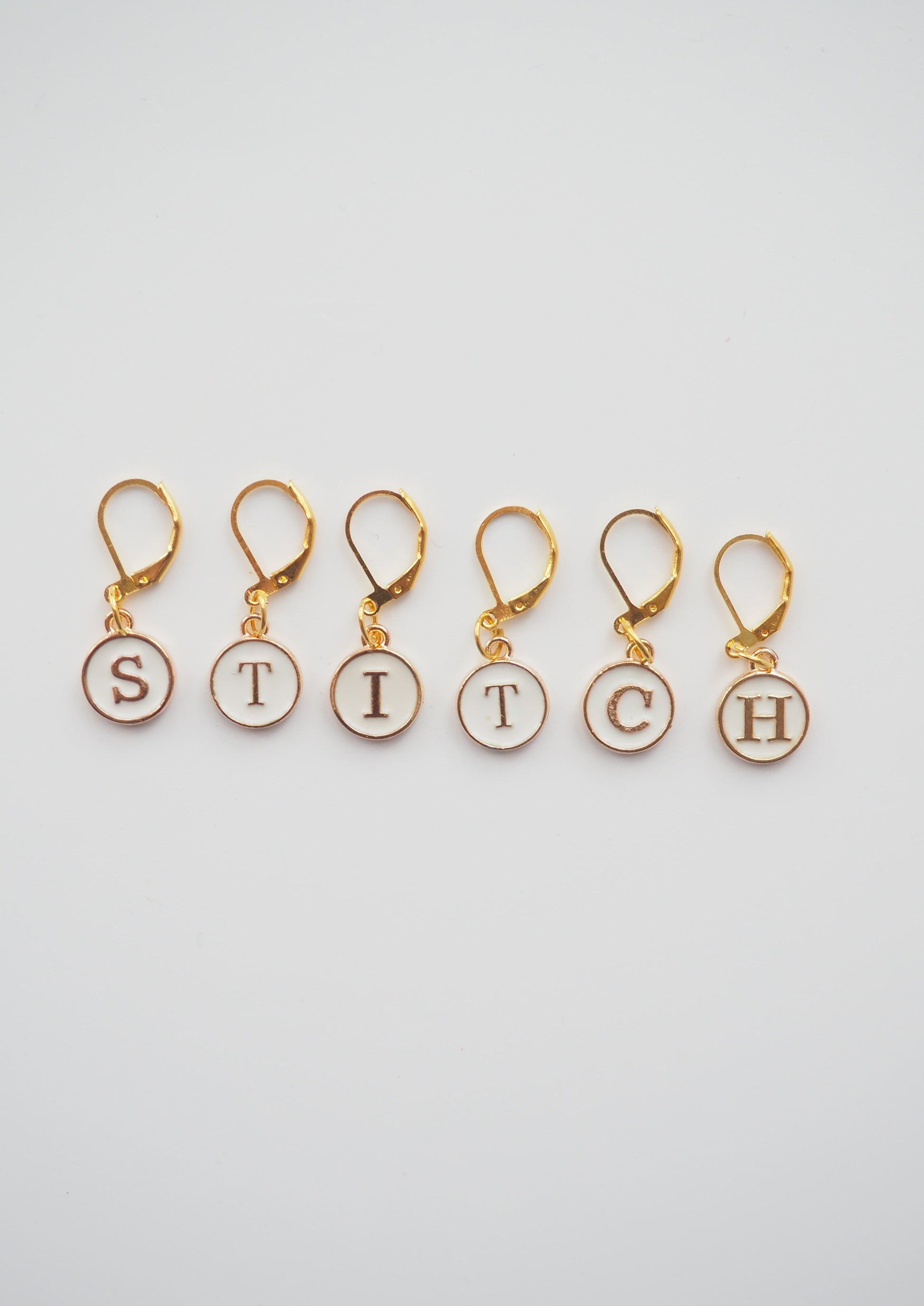 image of 6 white enamel stitch markers with gold colour metal. claw clasps which can be used for knitting or crochet. the markers spell out the word "stitch"