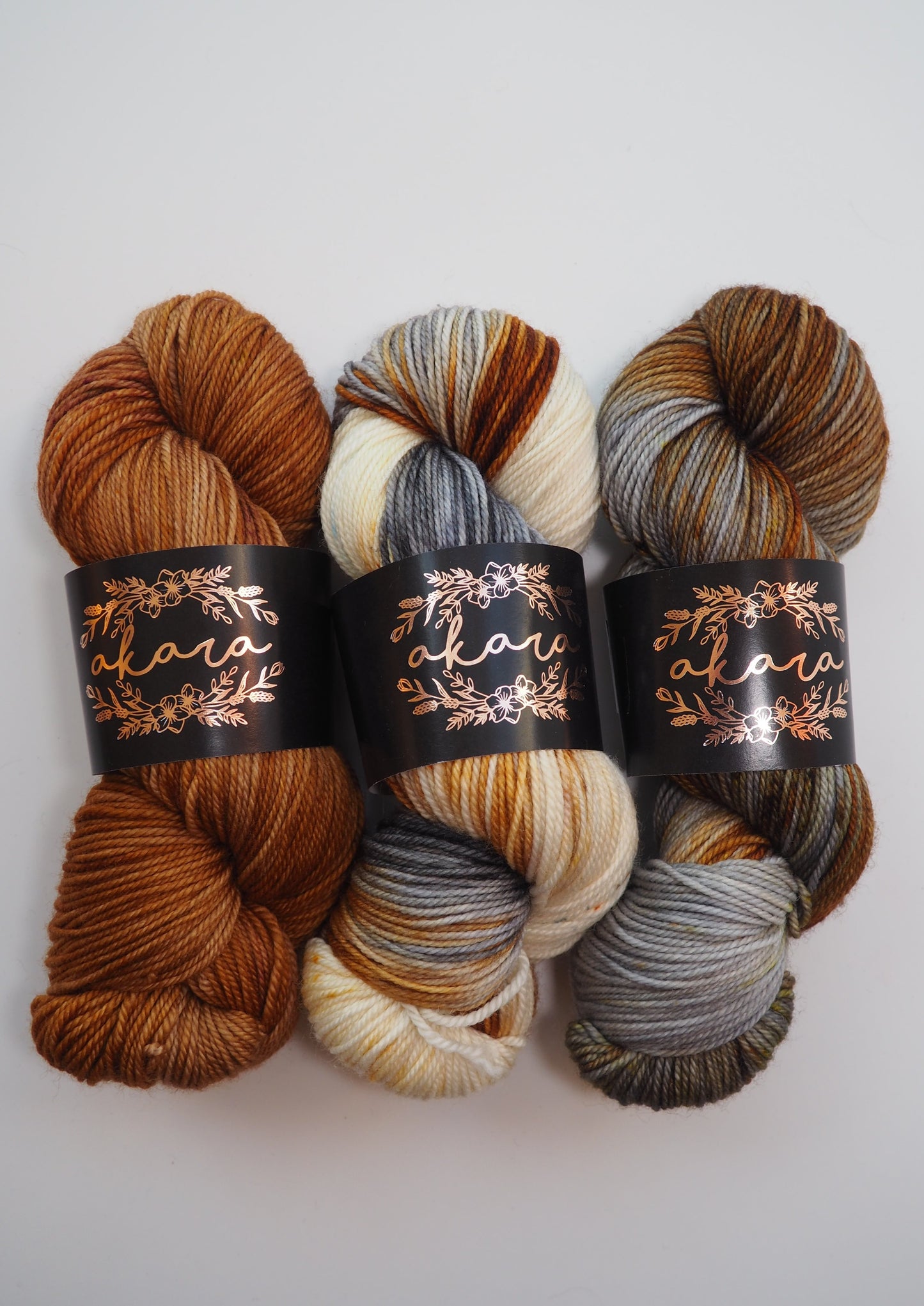 image of three skeins of akara merino dk, one in fallen leaves, one in bear with me (white, grey and brown), and likha (grey and brown)