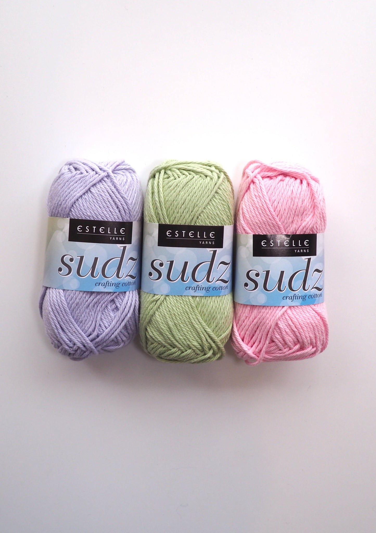 Image showcasing three balls of Estelle Cotton Sudz yarn. One ball is pistachio, one is violet, and one is pink. This versatile cotton yarn is perfect for durable crafts like washcloths and tote bags, as well as clothing items. Affordable and of high quality.
